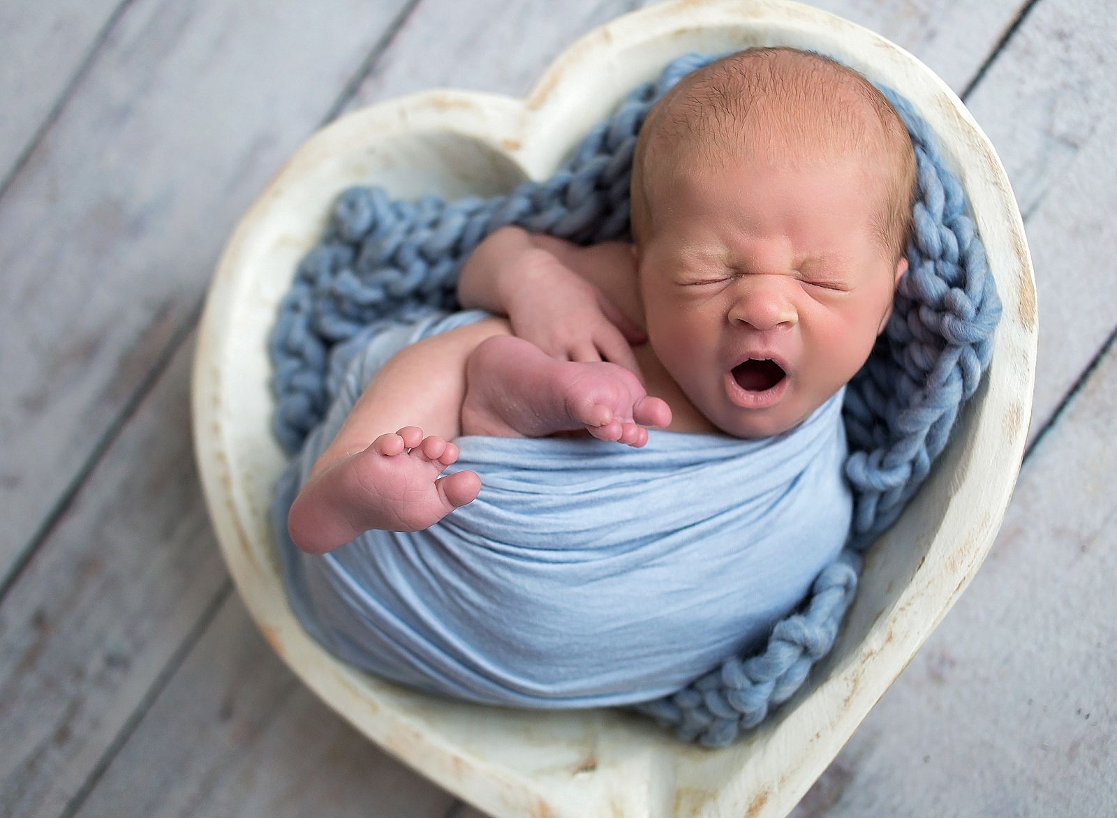 Newborn baby boy yawning while laying in a heart bowl wrapped in blue.
